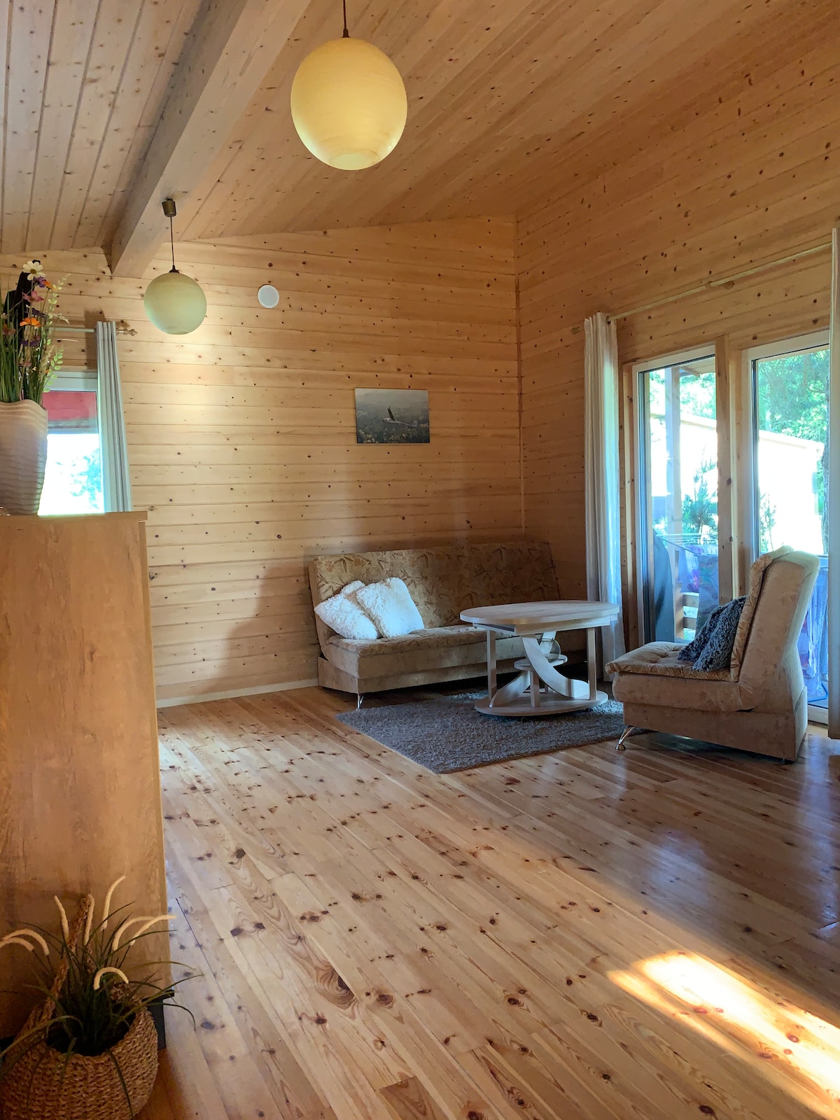 A cozy seating area features a plush sofa and an armchair placed beside a small table. Natural light streams through large glass doors, highlighting the wooden walls and floor. A decorative plant adds a touch of greenery to the space.