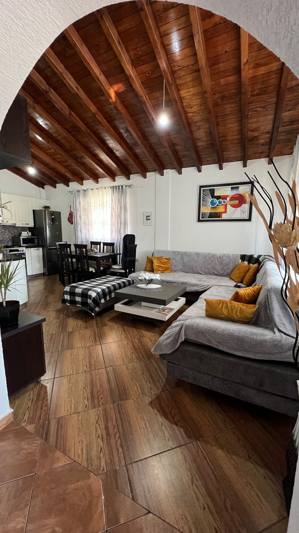 The living room features a spacious gray sectional sofa adorned with yellow accent pillows. A low coffee table is centered on a patterned rug, while a dining area with a dark table and chairs is visible in the background. Natural light filters through the curtains.