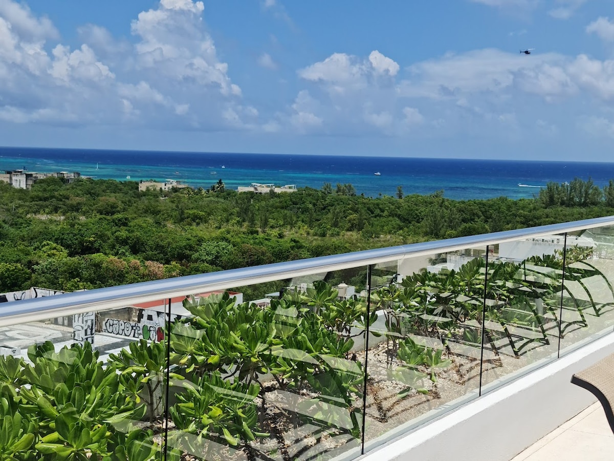 A panoramic view of the ocean from a rooftop terrace is presented, showcasing lush greenery in the foreground. The azure waters extend towards the horizon, where the sky meets the sea, illuminated by scattered clouds. The terrace railing features clear glass, enhancing the unobstructed vista.