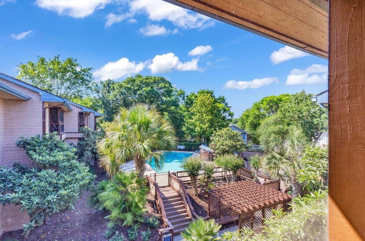 A view from the balcony showcases a lush, green landscape surrounding a clear blue pool. Wooden pathways and a pergola can be seen, inviting guests to explore the outdoor space and enjoy the tranquil setting. Tall trees provide shade, enhancing the serene environment.