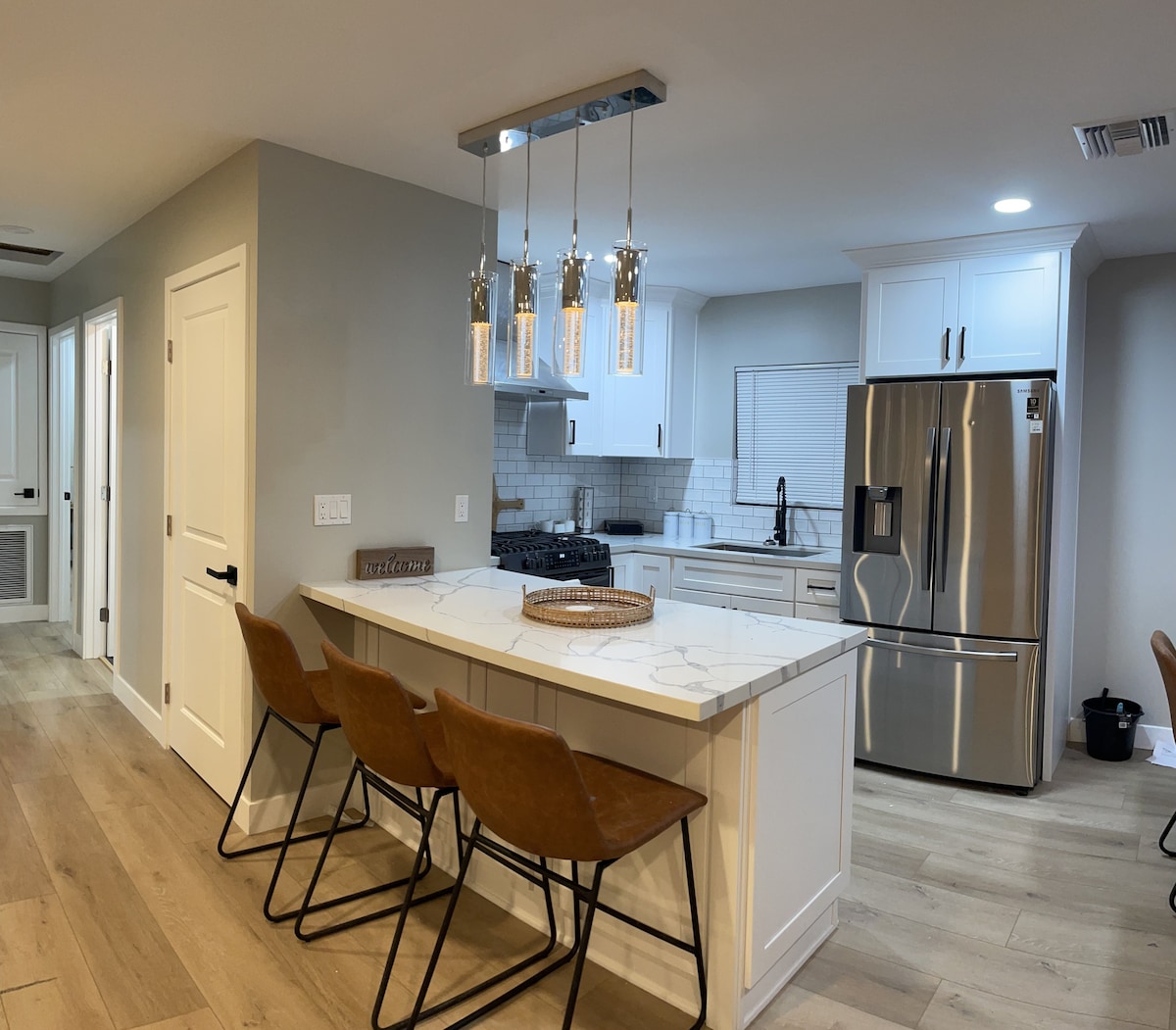 A modern kitchen features a white island with four brown leather stools arranged along one side. Stainless steel appliances, including a refrigerator, are positioned against the wall. Soft lighting is provided by hanging fixtures above the island, complementing the light wood flooring.