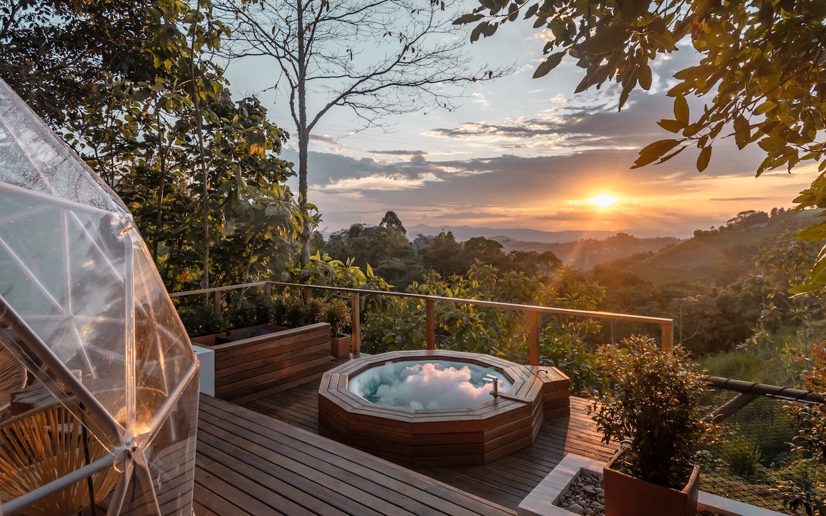 A wooden deck features a circular hot tub surrounded by lush greenery, with a stunning sunset in the background. The scene captures the tranquility of nature and offers a serene viewing experience of the landscape.