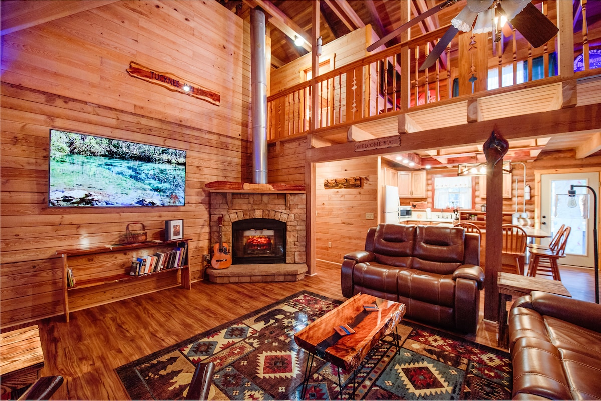 A spacious great room is highlighted by rich wooden walls and a cozy stone fireplace. A large flat-screen TV is mounted above the fireplace, while comfortable seating arrangements, a coffee table, and shelves filled with books are visible. A loft overlooks the area, adding to the cabin's open feel.