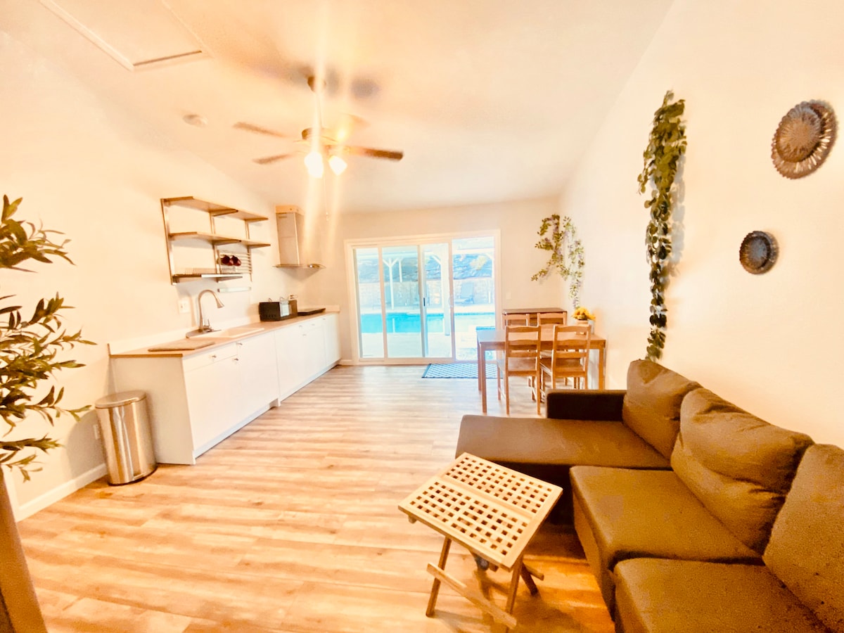 A bright living area is presented with a comfortable sofa and a small table. Shelving units are visible above the kitchenette, where appliances are neatly arranged. Natural light floods the space from large sliding doors leading to an outdoor area and pool.