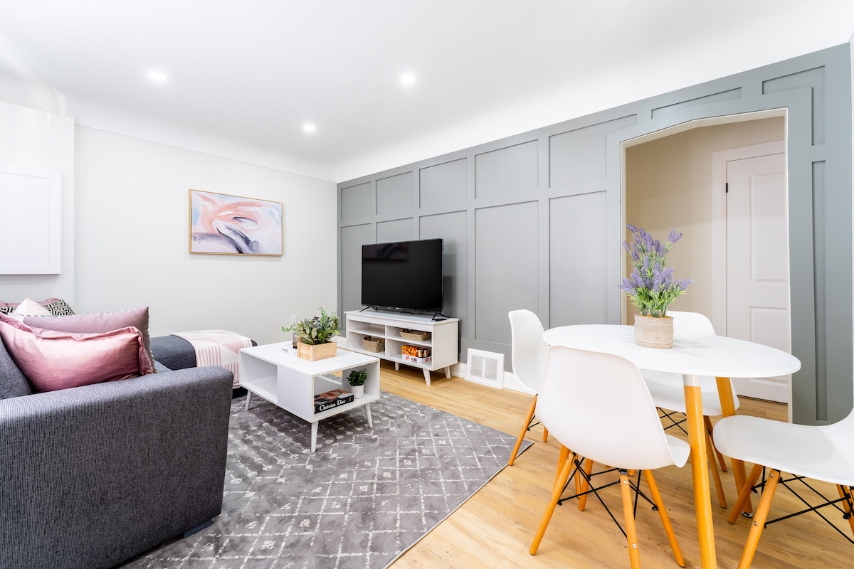 A modern living area is presented with a gray accent wall and stylish furnishings. A comfortable sofa is paired with a light wood coffee table, while a round dining table with white chairs is positioned nearby. A television sits on a media console, complemented by decorative plants.