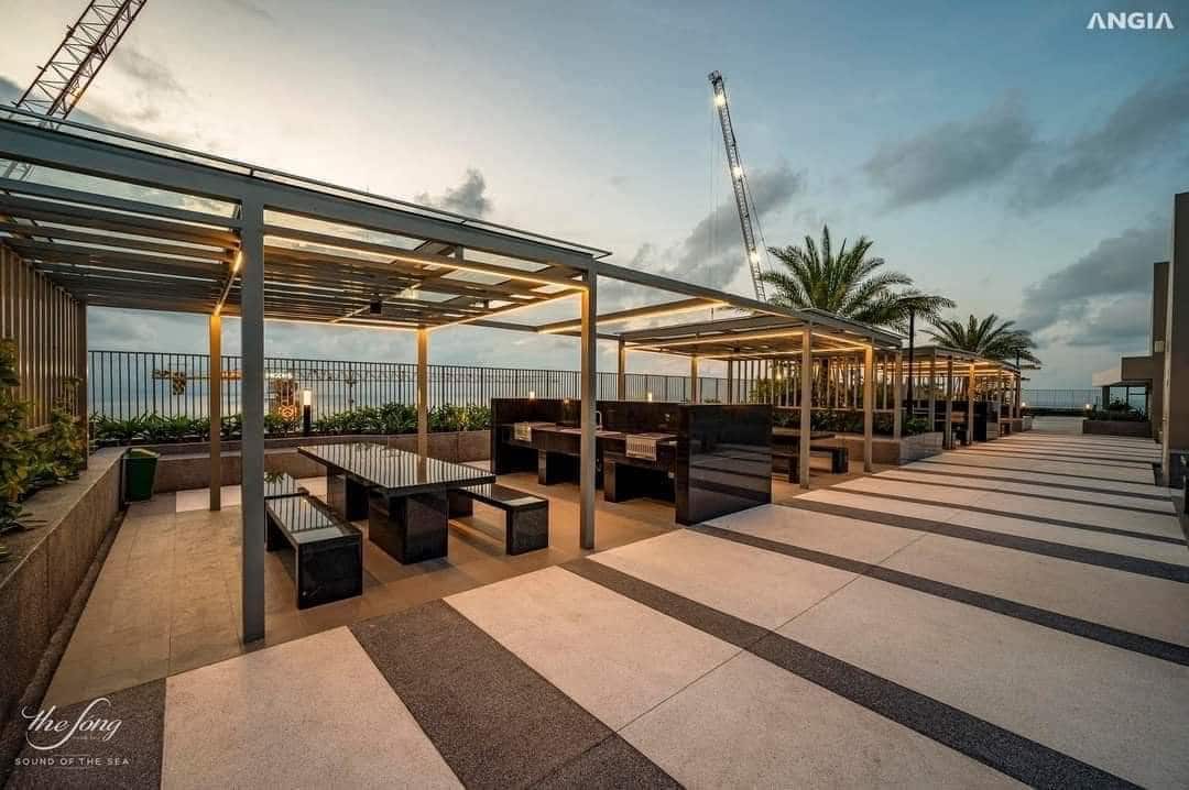 An outdoor dining area features several covered tables with black granite surfaces, providing ample seating. Lush greenery lines the perimeter, while palm trees add a tropical touch. The pathway is designed with large stone tiles, leading to the scenic views beyond.