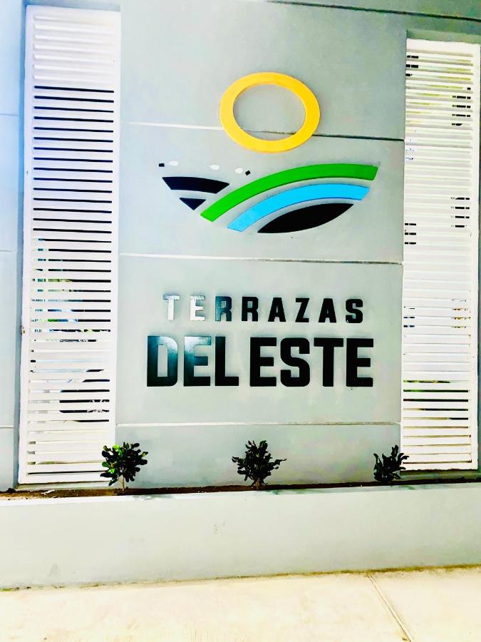 The entrance features a modern sign displaying the name 'Terrazas del Este' in bold letters. Colorful graphic elements suggest a connection to nature. Simple landscaping surrounds the sign, adding to the clean aesthetic of the peaceful neighborhood.