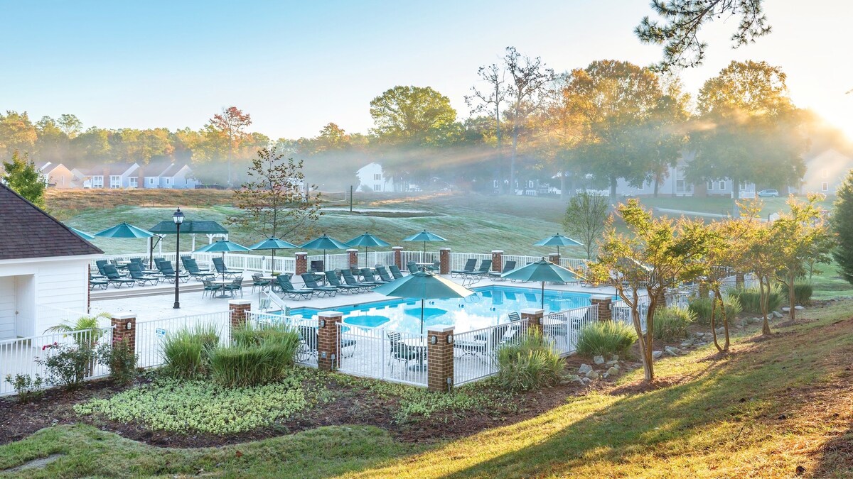 The outdoor pool area is surrounded by lounge chairs and umbrellas, set against a backdrop of lush greenery. Morning light filters through the trees, illuminating the space and creating a serene atmosphere for relaxation.