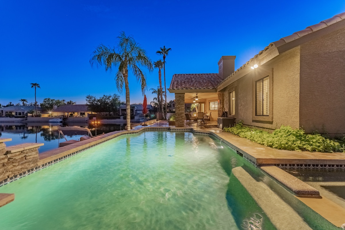 A serene pool area is captured at dusk, with water reflecting soft hues from the surrounding lights. Palm trees line the space, and a private dock can be seen in the distance, enhancing a tranquil atmosphere by the water's edge.