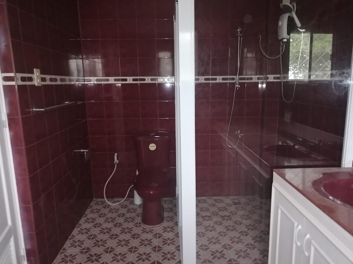 A bathroom is presented with deep red tiled walls and patterned floor tiles. A shower area is included, featuring a glass enclosure. A toilet is positioned in the corner, and a sink with a countertop completes the space, creating a cohesive design.