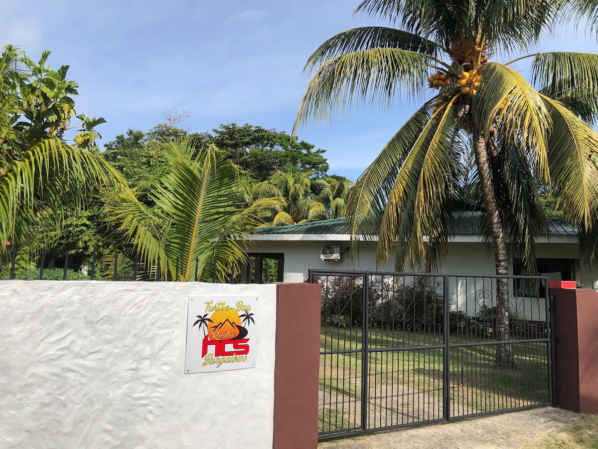 A welcoming entrance displays a sign for the property, surrounded by lush greenery and palm trees. The gated pathway leads towards a bungalow, partially obscured by foliage, contributing to a serene atmosphere. A well-maintained lawn is visible, enhancing the overall inviting look.