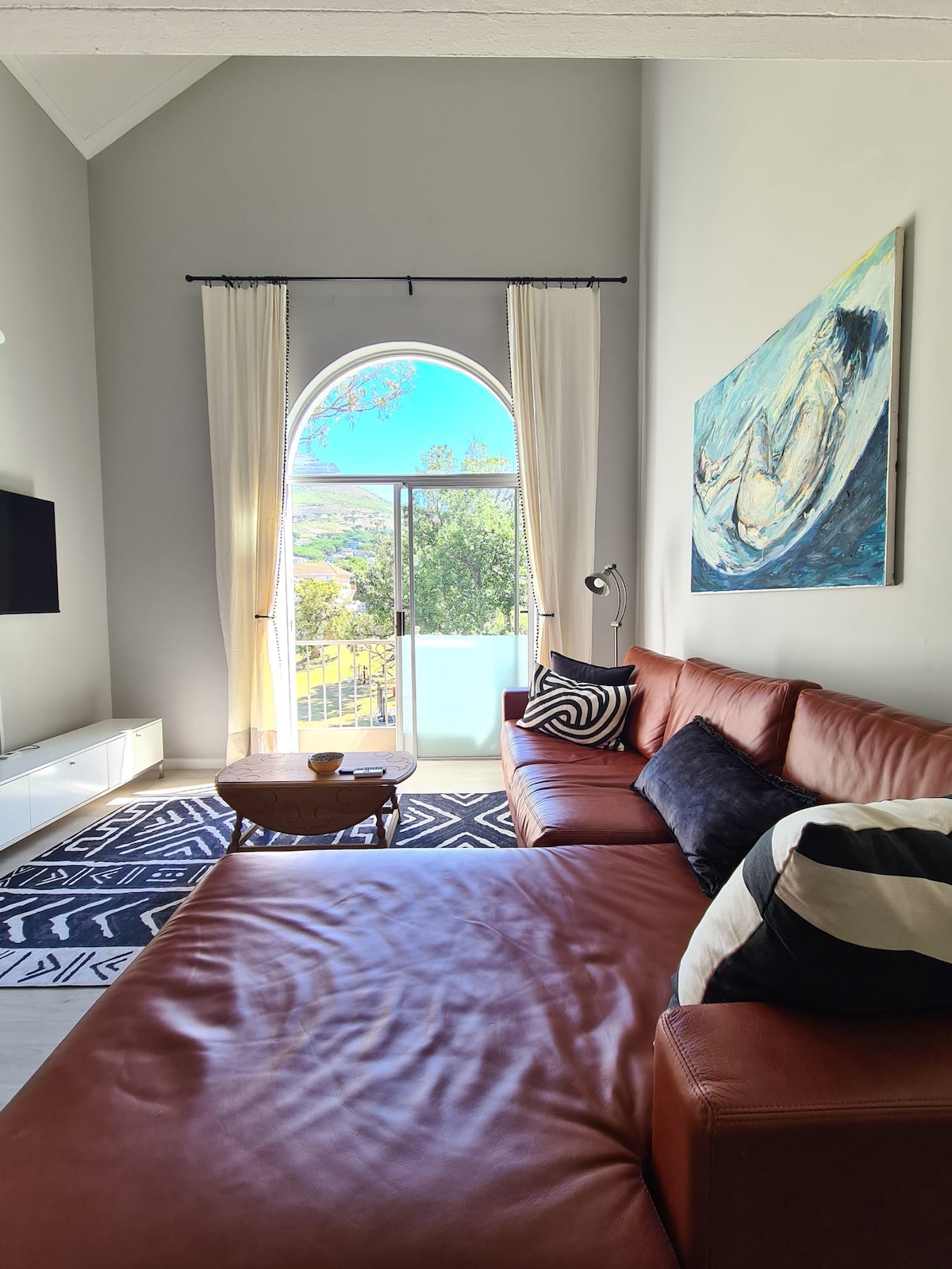 A spacious living area is highlighted by a large window framed with light curtains, offering views of the outdoors. A stylish leather sofa is paired with a round coffee table on a patterned area rug. An artwork adorns the wall, adding visual interest.