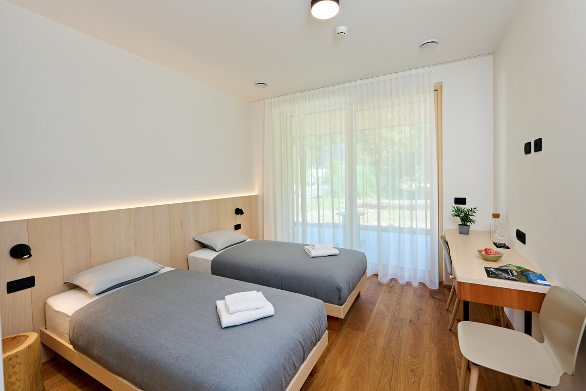 A bright and airy guest room is furnished with two single beds arranged side by side, each topped with a folded towel. Natural light filters through large sliding doors leading to a private terrace, while a small desk and chair are positioned against the wall.