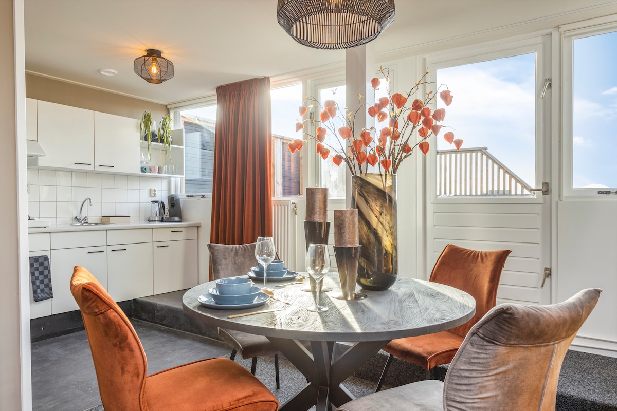 A dining area is presented with a round table set for four, featuring elegant dishware and glassware. Surrounding the table are four chairs in warm colors. Large windows allow natural light to fill the space, complemented by decorative branches in a vase.