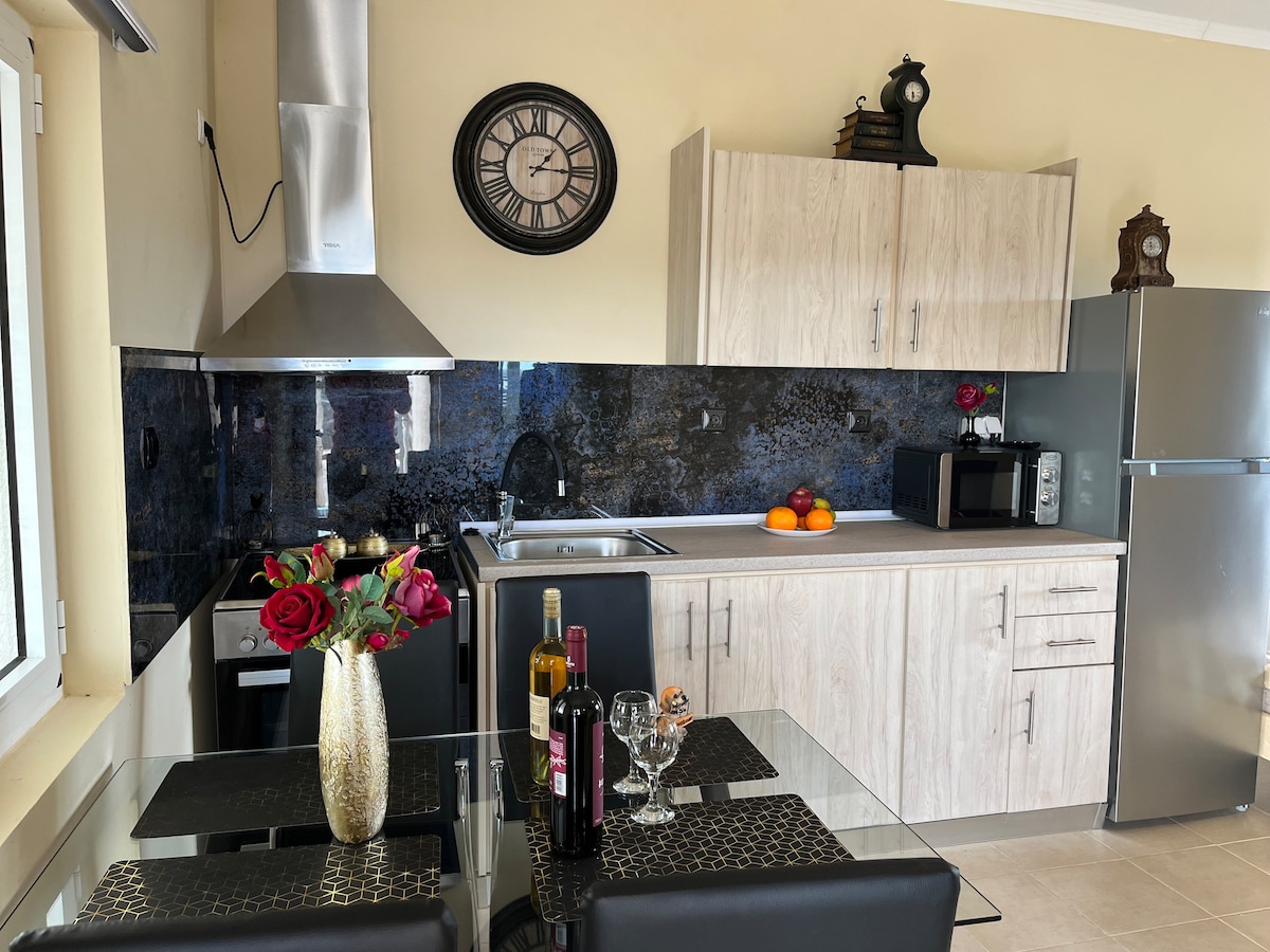A modern kitchen features sleek cabinetry and a marble-like backsplash. A dining table is set with wine glasses and bottles, accompanied by a vase of roses. Stainless steel appliances, including a microwave and refrigerator, are visible, while natural light enters through a nearby window.