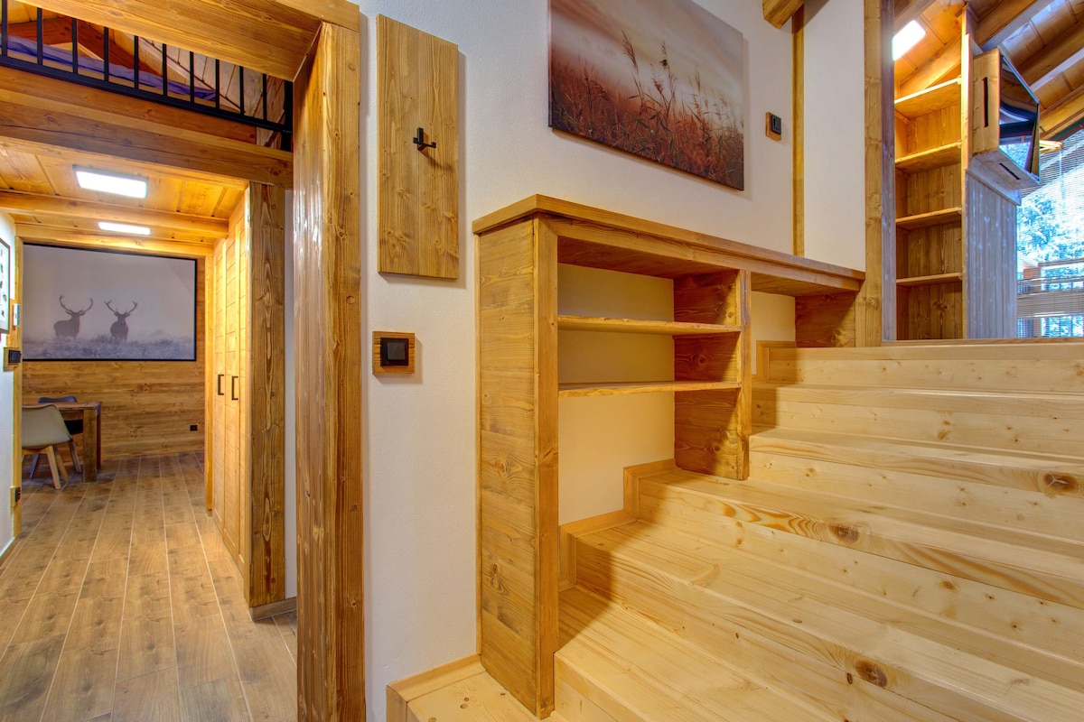 A wooden staircase is highlighted, leading to an inviting upper level. The walls feature natural wood paneling, and shelves are built into the staircase. Soft lighting illuminates the space, enhancing the warm tones of the wood elements.