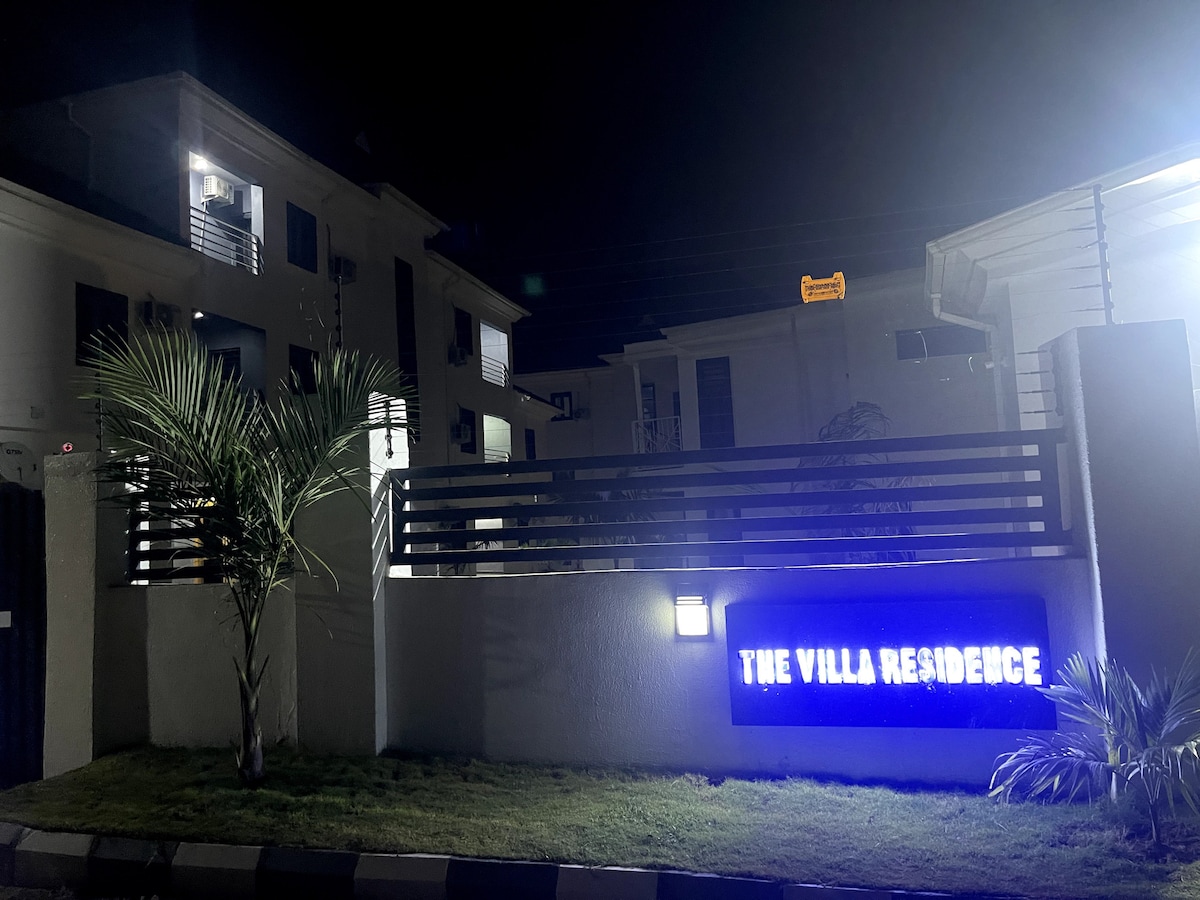 The exterior of The Villa Residence is illuminated at night, featuring a modern entrance highlighted by a glowing sign. Lush greenery is observed in the foreground, complemented by a well-maintained lawn that enhances the property’s appeal.