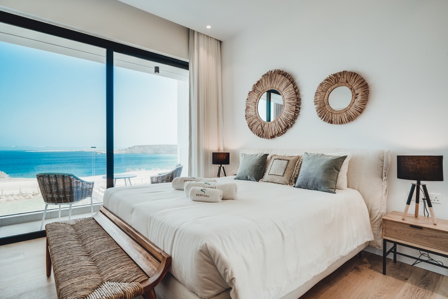 A bright bedroom features a spacious bed with neutral linens positioned in front of large glass doors, offering views of the sea. Two circular mirrors adorn the wall, while bedside tables with lamps are placed on either side. A comfortable seating area is visible outside.