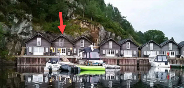 A row of wooden buildings sits along a dock, reflecting on calm waters. Each structure has a sloped roof and large windows. A kayak and two motorboats are positioned in front, with a rocky hillside providing a natural backdrop.