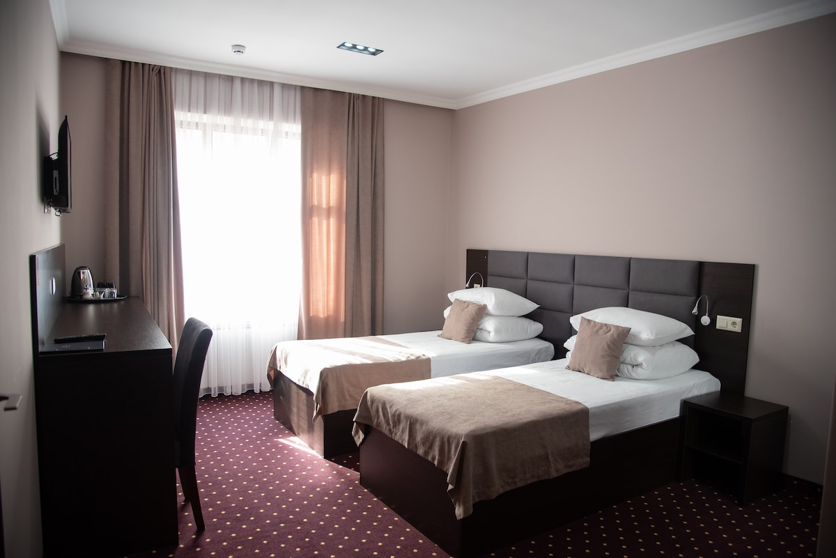 The room features two spacious beds with neutral-colored linens and decorative pillows. A desk and chair are positioned against the wall, with a small television mounted above. Natural light enters through a large window covered by sheer curtains, complementing the warm color scheme.