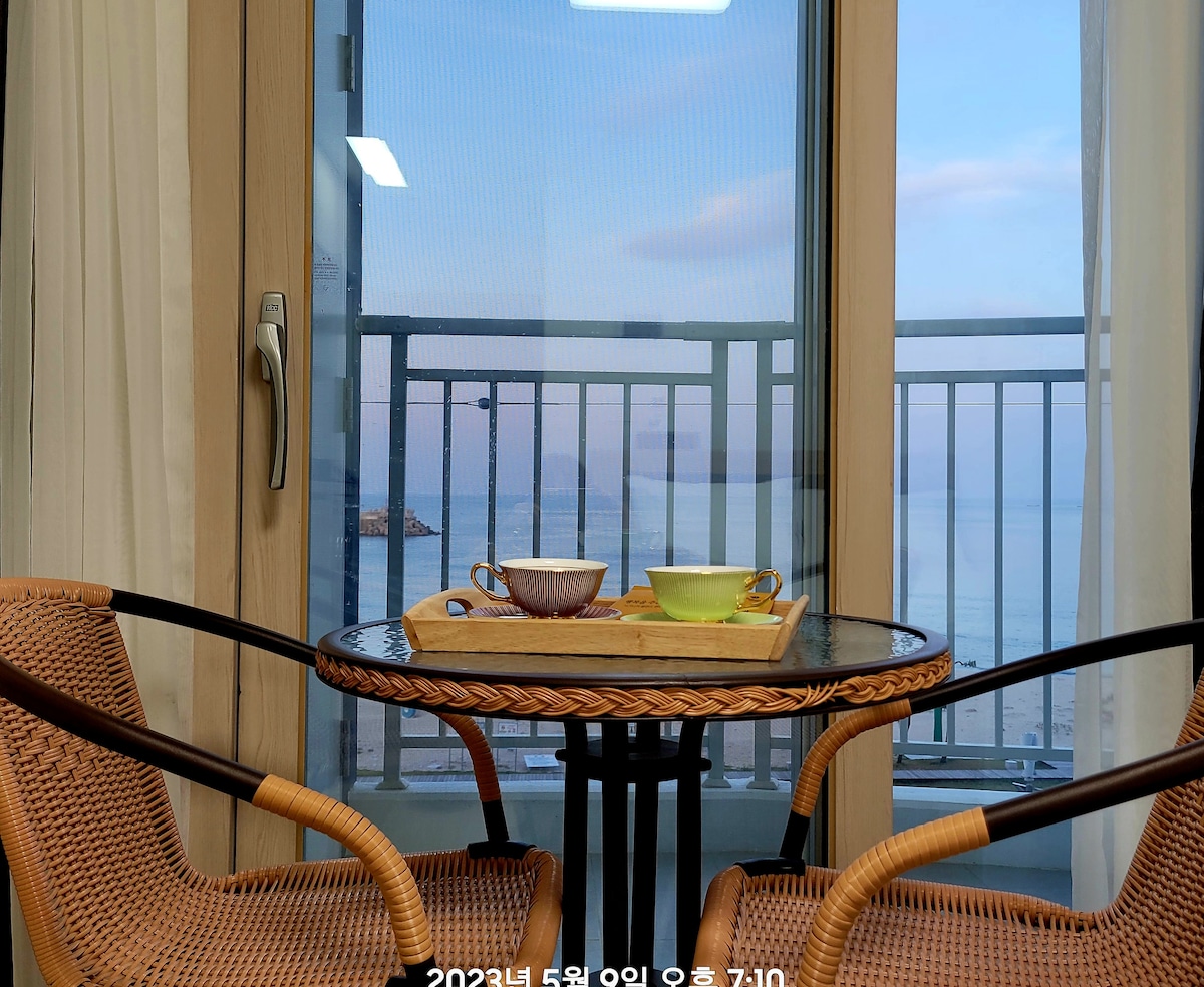 A cozy breakfast setting is visible with a round table topped with a wooden tray holding two colorful bowls. The table is surrounded by wicker chairs, and through the large window, a serene view of the ocean can be seen, reflecting soft morning light.