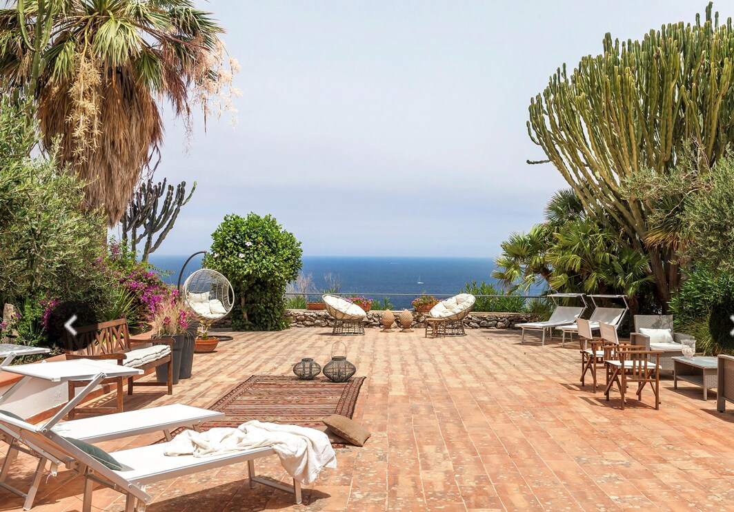A spacious outdoor terrace is presented, featuring various seating arrangements and sun loungers, surrounded by lush greenery and colorful plants. An expansive view of the sea is visible in the background, creating a serene setting for relaxation and dining.