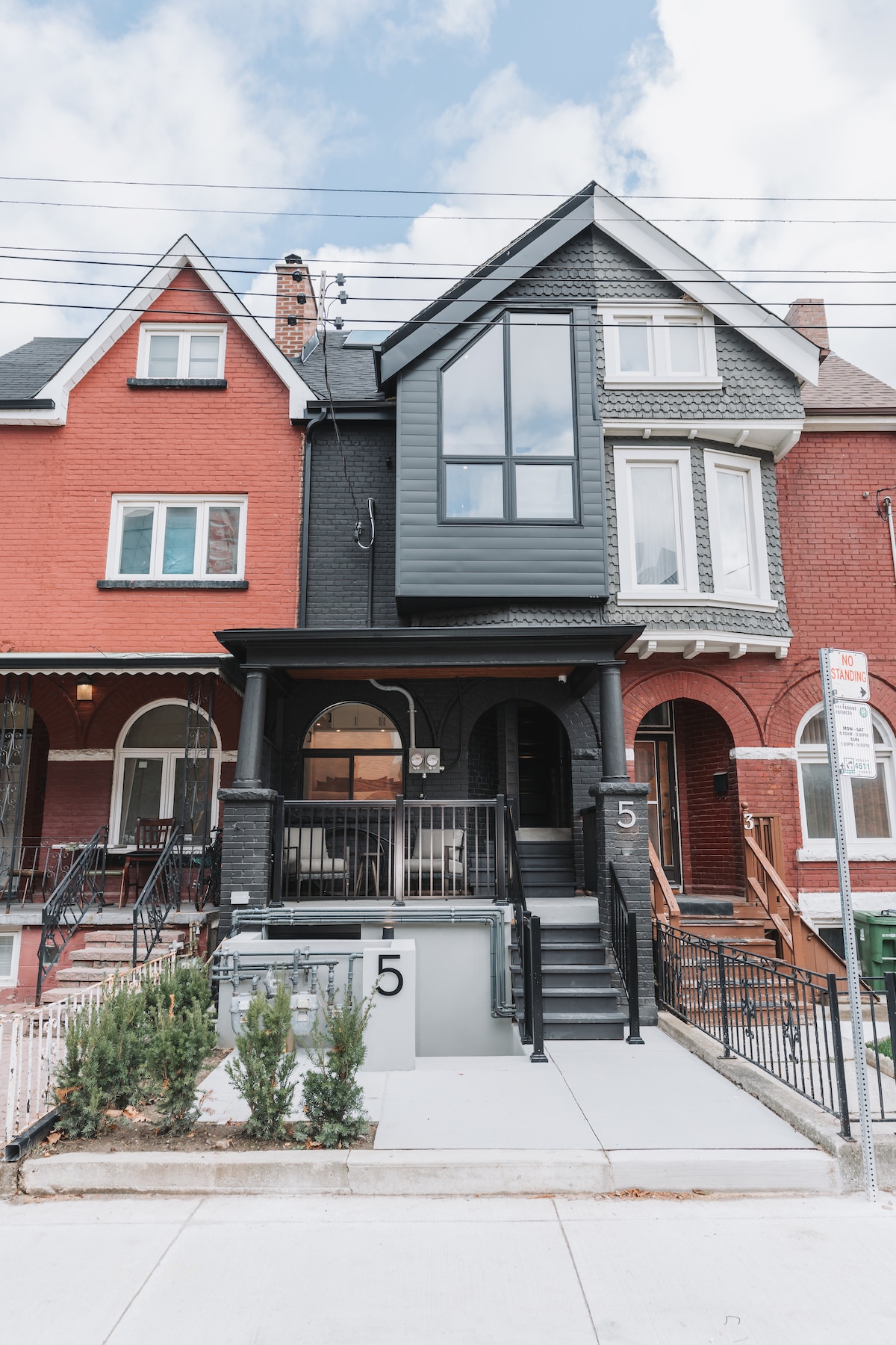 A sleek, modern triplex exterior is captured, featuring a dark facade and large windows. The entrance is highlighted by a set of steps leading to a welcoming front porch, while manicured greenery lines the pathway, blending urban living with a touch of nature.