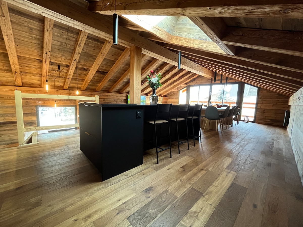 An open kitchen and dining area feature wooden beams and warm wood flooring. A large island bar with stools invites gatherings, while a spacious dining table is illuminated by natural light from expansive windows. Minimalist decor enhances the inviting atmosphere.