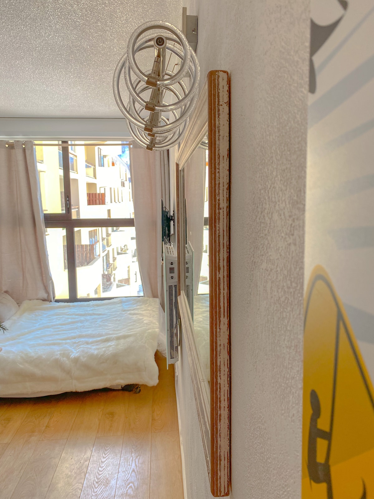 A cozy corner of the studio features a wall-mounted mirror framed in distressed wood. A light fixture with circular design hangs above, while a spacious bed is visible in the background, draped with a soft white blanket. Natural light filters through the large windows.