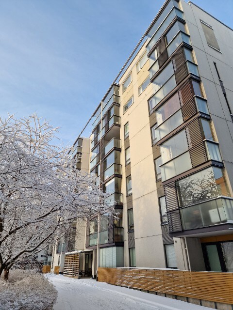 The exterior of a modern apartment building is shown, featuring multiple balconies and large glass windows. Fresh snow covers the ground and tree branches, creating a serene winter landscape. The building's design includes a blend of light-colored walls and wooden accents.