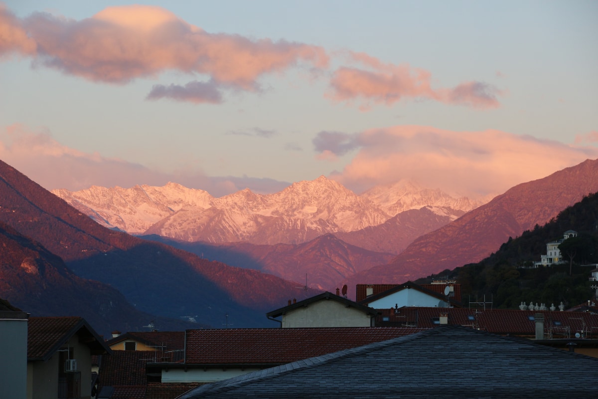A mountain landscape is captured, featuring snow-capped peaks bathed in soft pink light. The foreground showcases rooftops of nearby buildings, contrasting with the natural beauty of the surrounding mountains and sky.