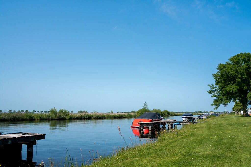 A serene canal is lined with boats, resting on wooden docks. Lush green grass stretches along the water's edge, while tall grasses and trees frame the scene. The bright blue sky enhances the tranquil atmosphere of this natural setting.