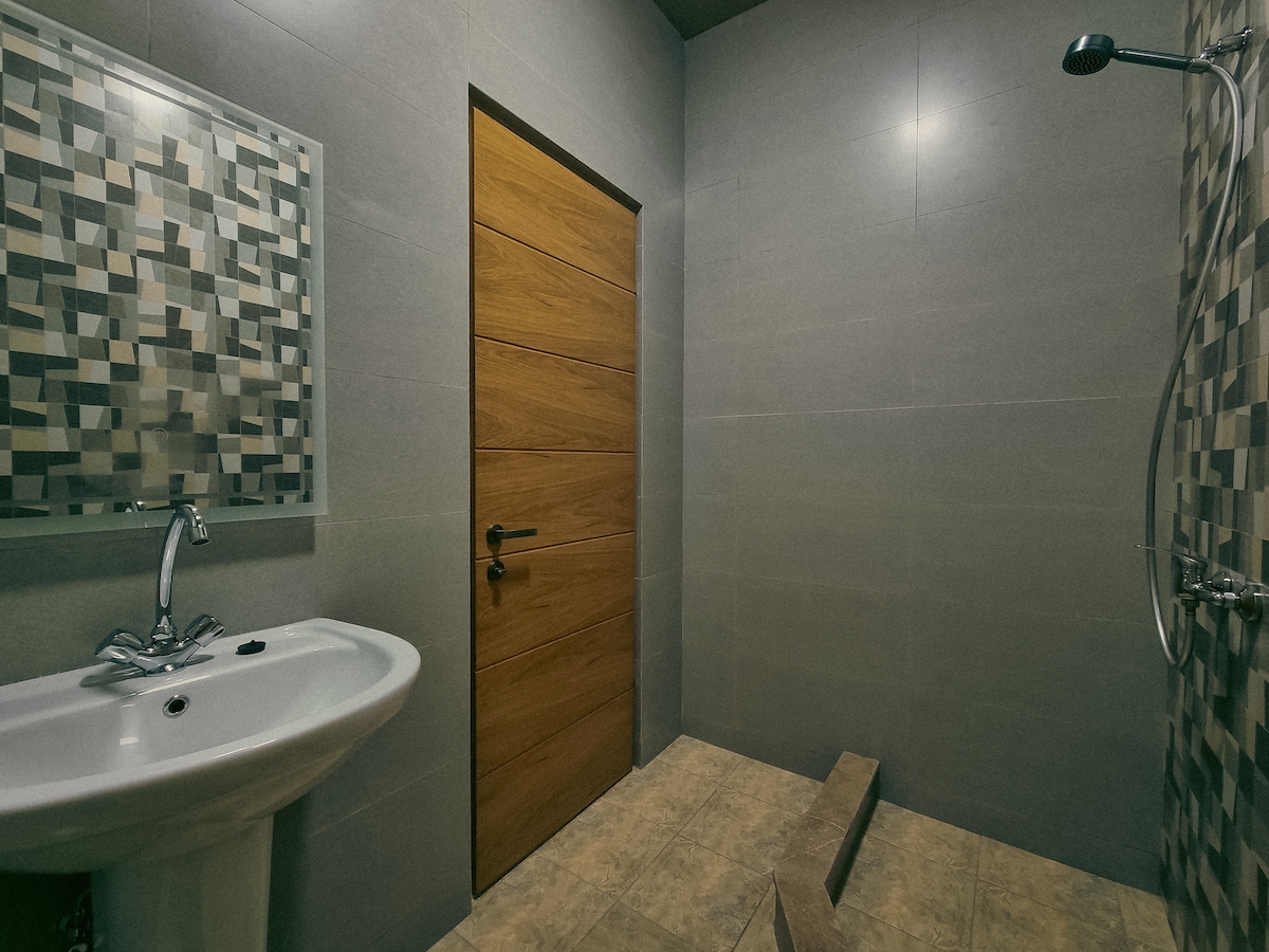 A modern bathroom features a sleek sink with a wall-mounted faucet and a spacious walk-in shower. Textured tile accents are present on one wall, while a wooden door provides access to the space. Neutral tones create a clean and calming environment.