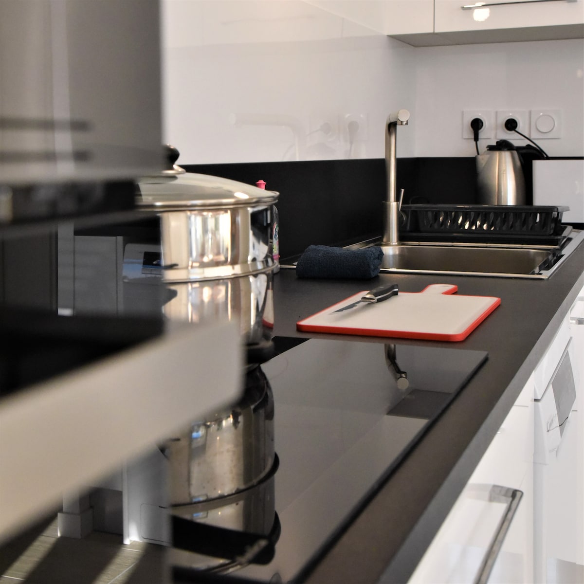 A modern kitchen area features sleek black countertops and a reflective surface. Kitchen appliances, including a kettle and a microwave, are visible, along with a stainless steel pot. A cutting board sits on the counter, accompanied by a dish drying rack and a cloth.