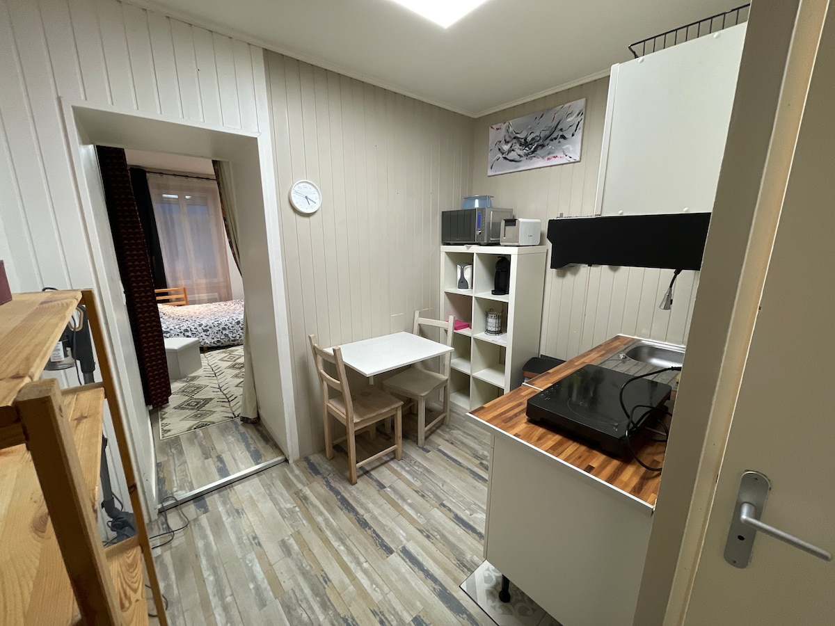 The kitchen area is equipped with a small table and two chairs. A modern black stove is positioned on a wooden counter. Shelves displaying kitchen essentials are present, while a corridor leads toward a bedroom area, partially visible in the background.