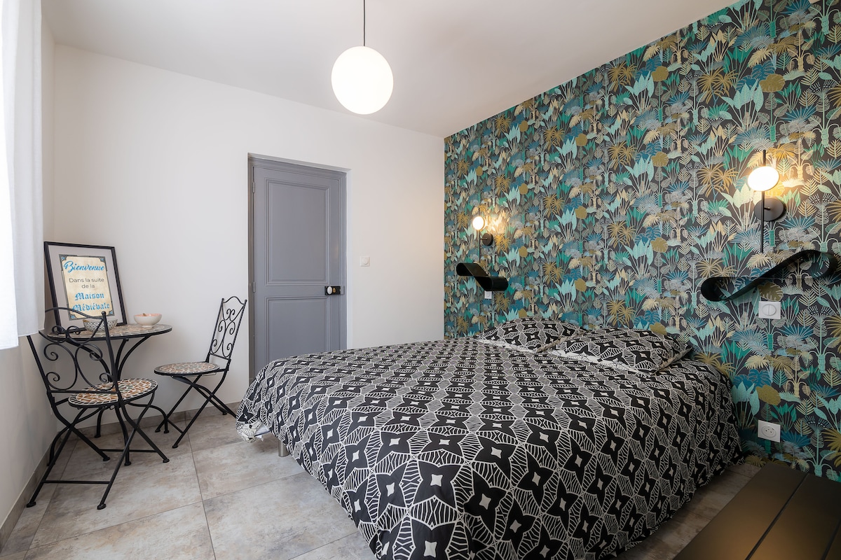 A welcoming bedroom features a bold patterned accent wall, complemented by soft lighting. A spacious bed is adorned with a geometric patterned cover. A small table and two chairs are positioned nearby, enhancing the cozy ambiance of the room.