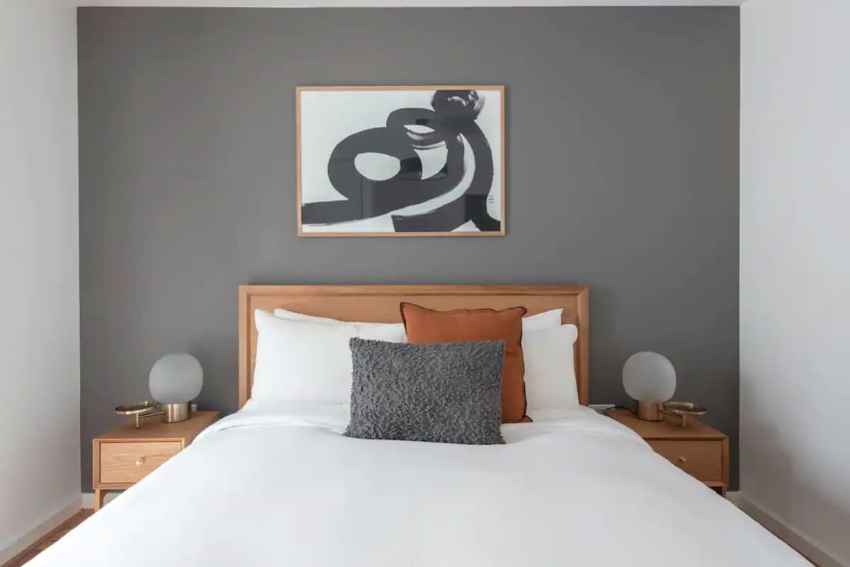 The bedroom features a spacious bed with a light-colored duvet and an arrangement of pillows. A decorative abstract artwork is displayed above the headboard. Two bedside tables with round lamps create a balanced aesthetic against the gray accent wall.