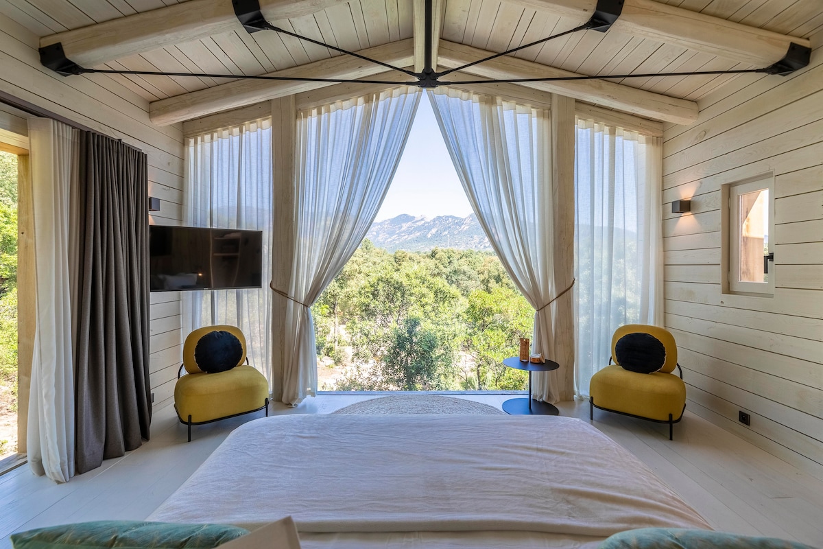 A spacious bedroom features a large bed with soft linens and expansive glass doors that open to stunning mountain views. Natural light is welcomed through sheer curtains, while two yellow accent chairs and a small side table complete the inviting layout.