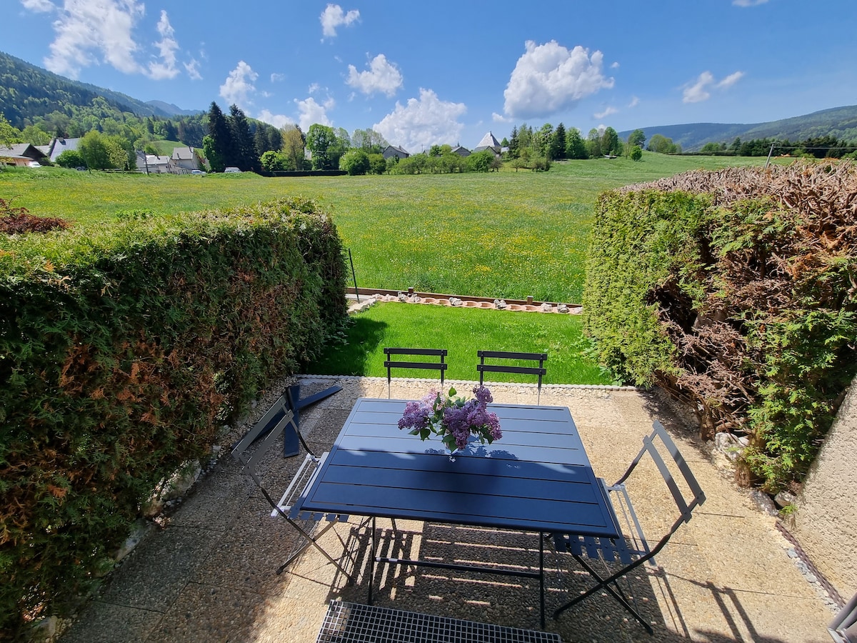 A terrace area is visible with a black outdoor dining table and chairs, surrounded by well-maintained hedges. A vase with flowers is placed on the table. In the background, an expansive grassy field and distant hills are framed by a clear blue sky with fluffy clouds.
