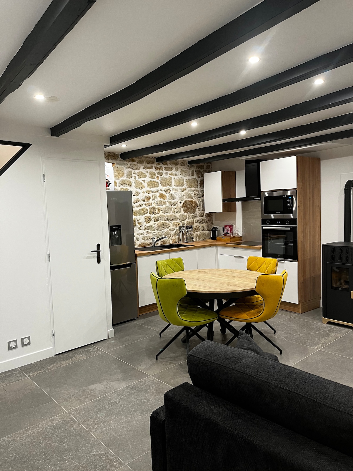A modern kitchen features exposed stone walls, complemented by sleek cabinetry and stainless steel appliances. A round wooden table is surrounded by four chairs in bright yellow and green hues. Soft lighting illuminates the space, creating an inviting atmosphere.