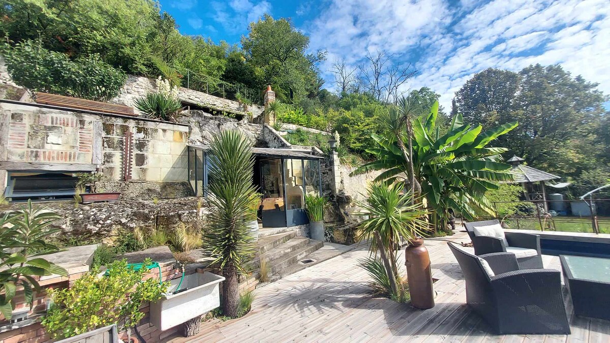The exterior area features a wooden deck surrounded by lush greenery and unique plant arrangements. The hillside residence is visible in the background, showcasing natural stone walls and large glass doors that open to the interior. Comfortable outdoor seating invites relaxation.