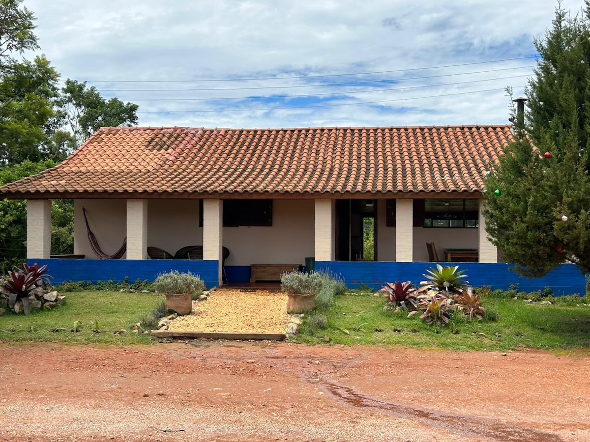 A single-story house features a traditional tile roof and wooden accents. A porch with seating options is highlighted, surrounded by a landscaped garden with various plants and flowers. Natural light filters through the windows, providing an inviting view of the outdoors.
