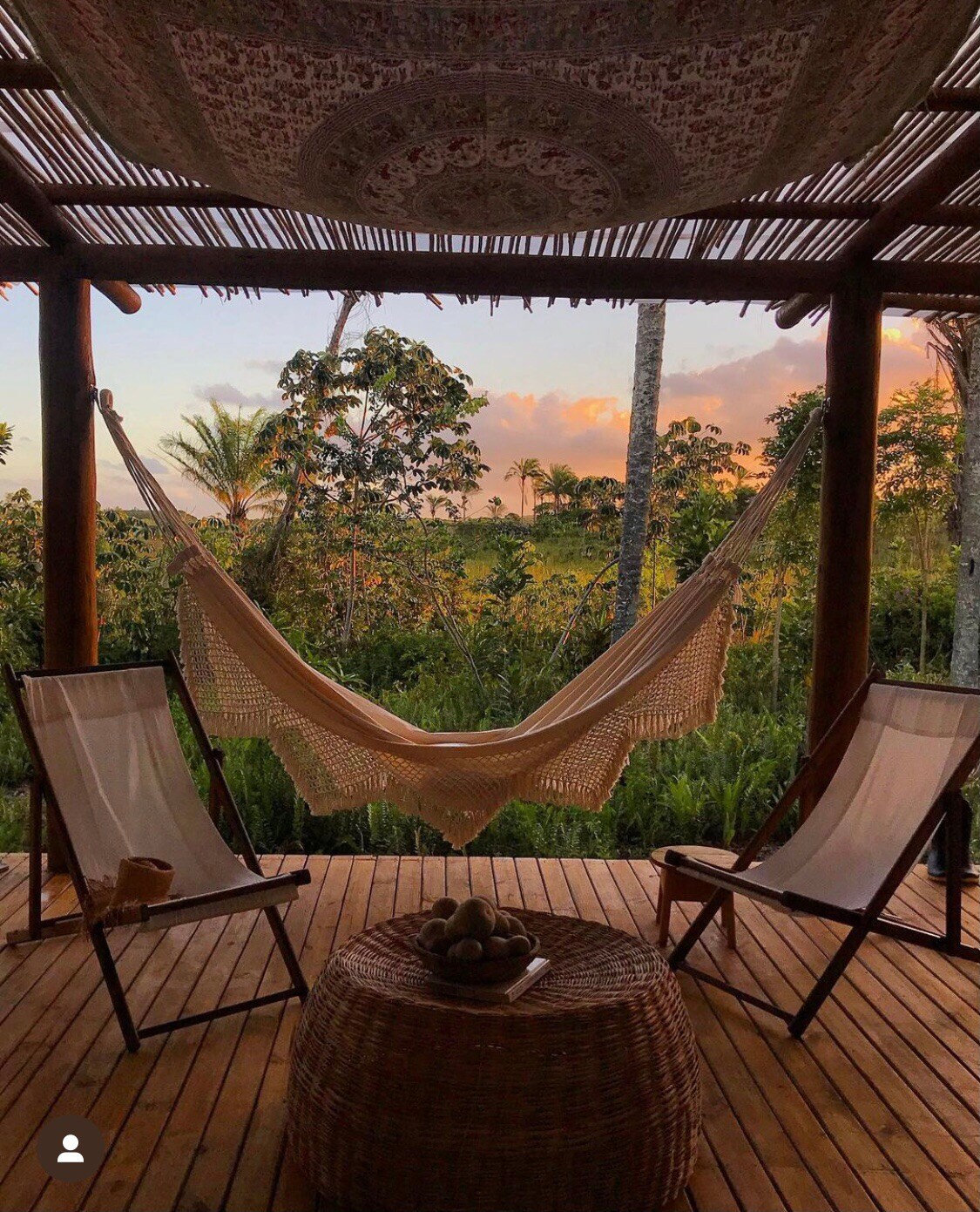 A cozy outdoor space features two wooden chairs positioned beside a round wicker table. A lightly woven hammock hangs between two posts, offering a view of lush greenery and a colorful sky at sunset. Natural light filters through the open beams above.
