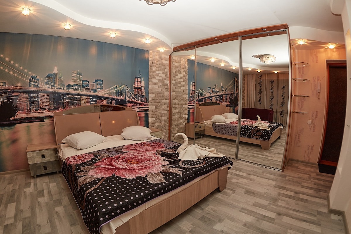 A cozy bedroom features a large mirror across from a spacious bed adorned with a floral patterned blanket. The walls display a city skyline mural, illuminated by soft lighting fixtures on the ceiling, while a neutral-toned floor adds a calming touch.
