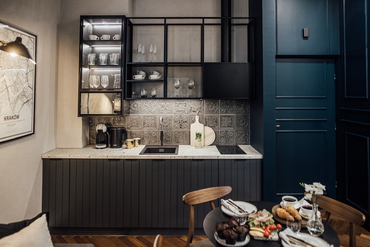 The kitchen area features a modern design with dark cabinetry and a light marble countertop. Glassware is neatly arranged on open shelving. A coffee machine and essential appliances are visible. A dining table is set with plates, cutlery, and a selection of baked goods.