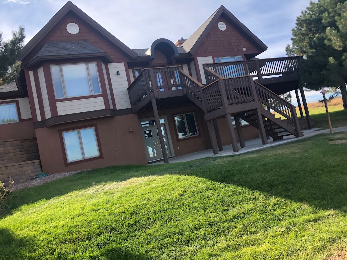 A two-story house features a welcoming exterior with a combination of brown and cream siding. A set of stairs leads to a spacious upper deck, surrounded by green grass and trees. Large windows are present, allowing natural light to enhance the façade.