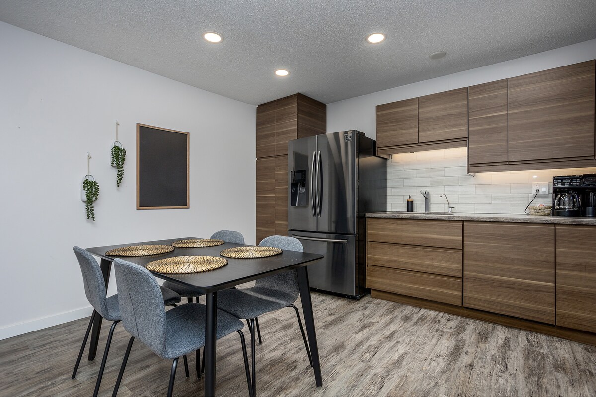 A modern kitchen area features sleek wooden cabinetry and stainless steel appliances. A dining table with four grey chairs is positioned centrally, accompanied by woven placemats. Soft overhead lighting illuminates the space, enhancing the minimalist design.