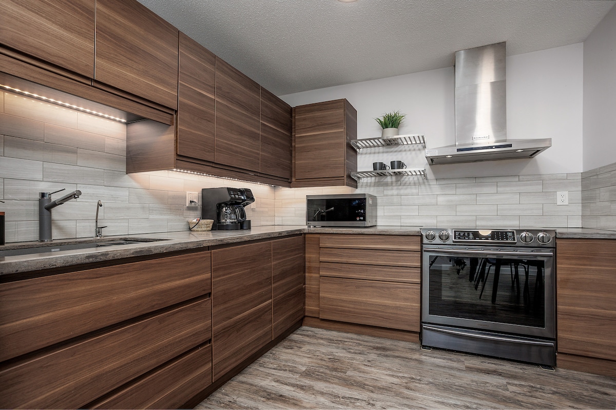 The kitchen features modern, wooden cabinetry with ample storage space. Stainless steel appliances, including an oven and microwave, are integrated into the design. A coffee maker is visible, and soft lighting illuminates the countertops, enhancing functionality in the cooking area.