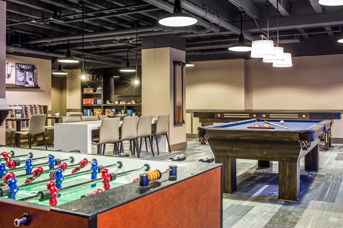 An entertainment area features a foosball table and a billiards table, inviting relaxation and socialization. Surrounding seating areas with tables are available for games or gatherings. Soft lighting from overhead fixtures creates a comfortable environment for guests to enjoy leisure activities.