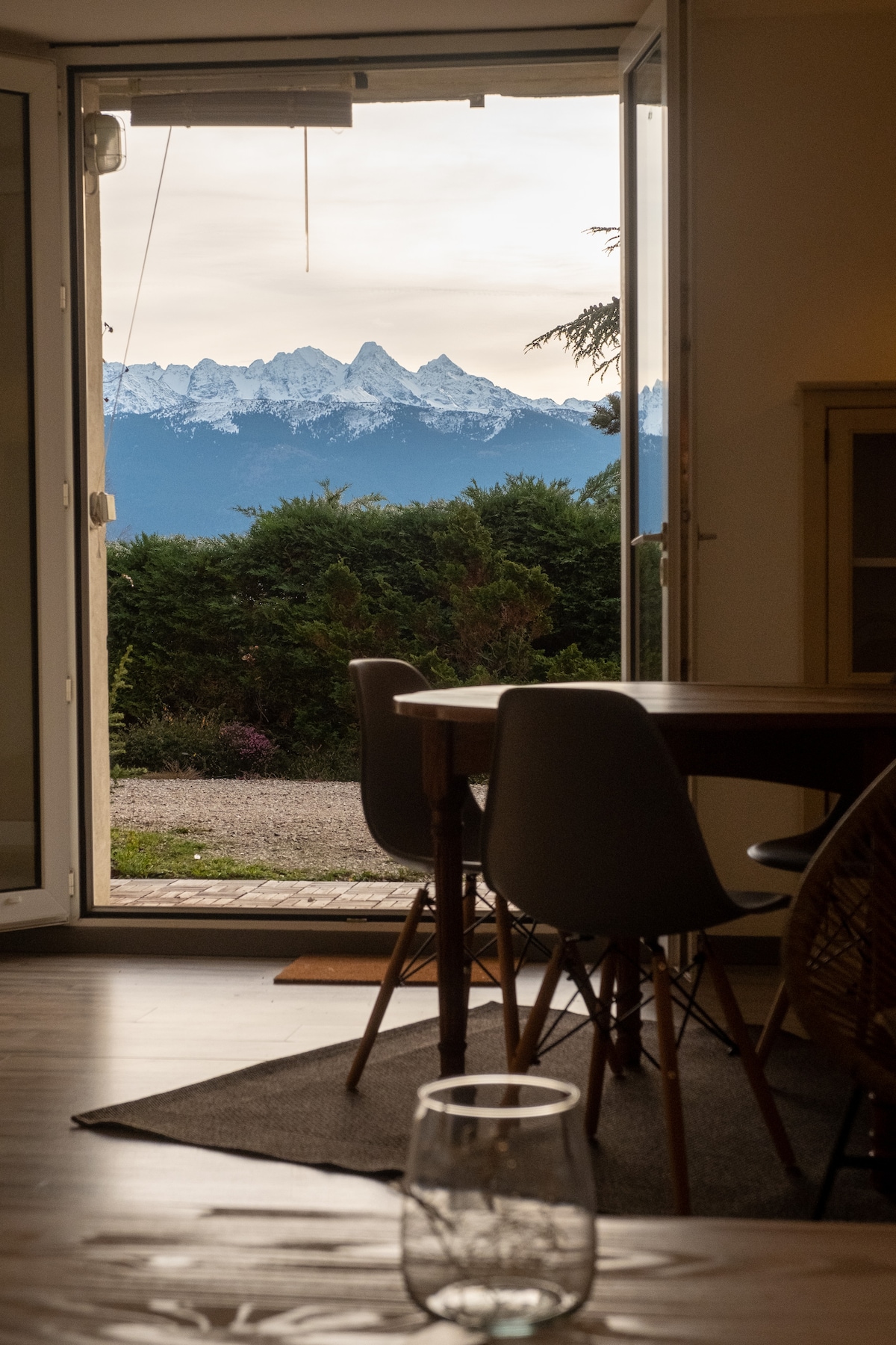 A view of snow-capped mountains is framed by an open door, offering a glimpse into a welcoming interior space. A round dining table with modern chairs is visible, alongside a partial view of a glass resting on a smooth surface, creating a serene atmosphere.